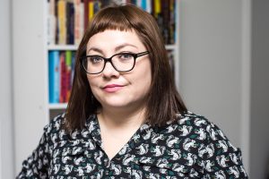 Isabel Quintero headshot. Isabel has straight, shoulder-length auburn hair with bangs and wears glasses and a black and white button-up shirt in front of bookshelves.