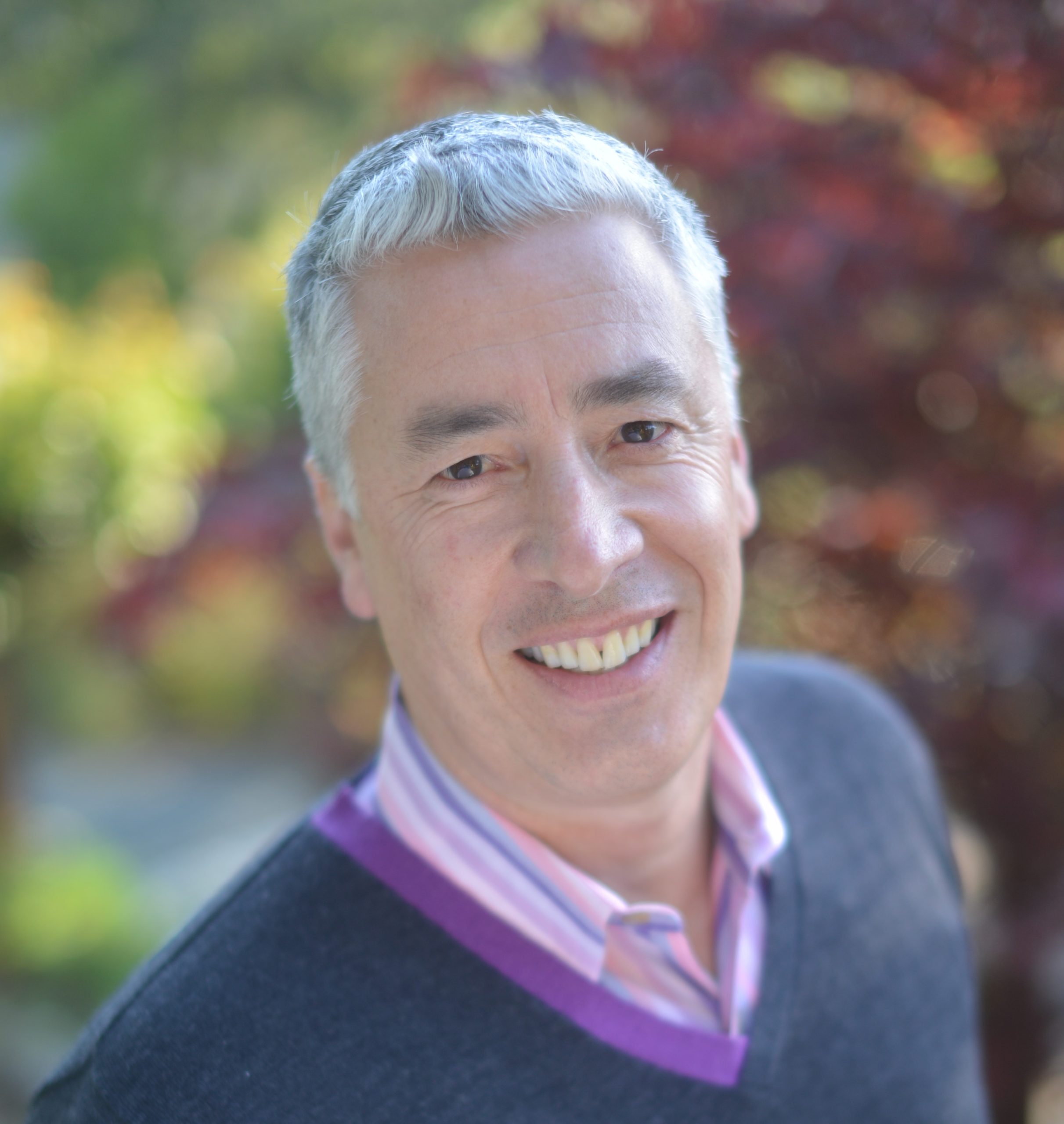 Michael Genhart author photo: Michael has short, gray hair, wears a pink collared shirt under a navy sweater, and stands in front of a leafy background.